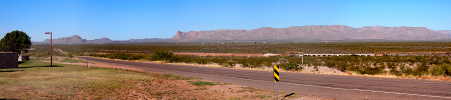 [A long, flat expanse of ground with lots of low-growing sagebrush. In the distance buildings sit near the edge of a sandstone-colored mountain range.]